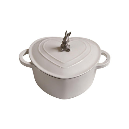 A heart-shaped white enameled cast iron pot with side handles and a small metallic rabbit figurine on top of the lid, shown against a plain white background.