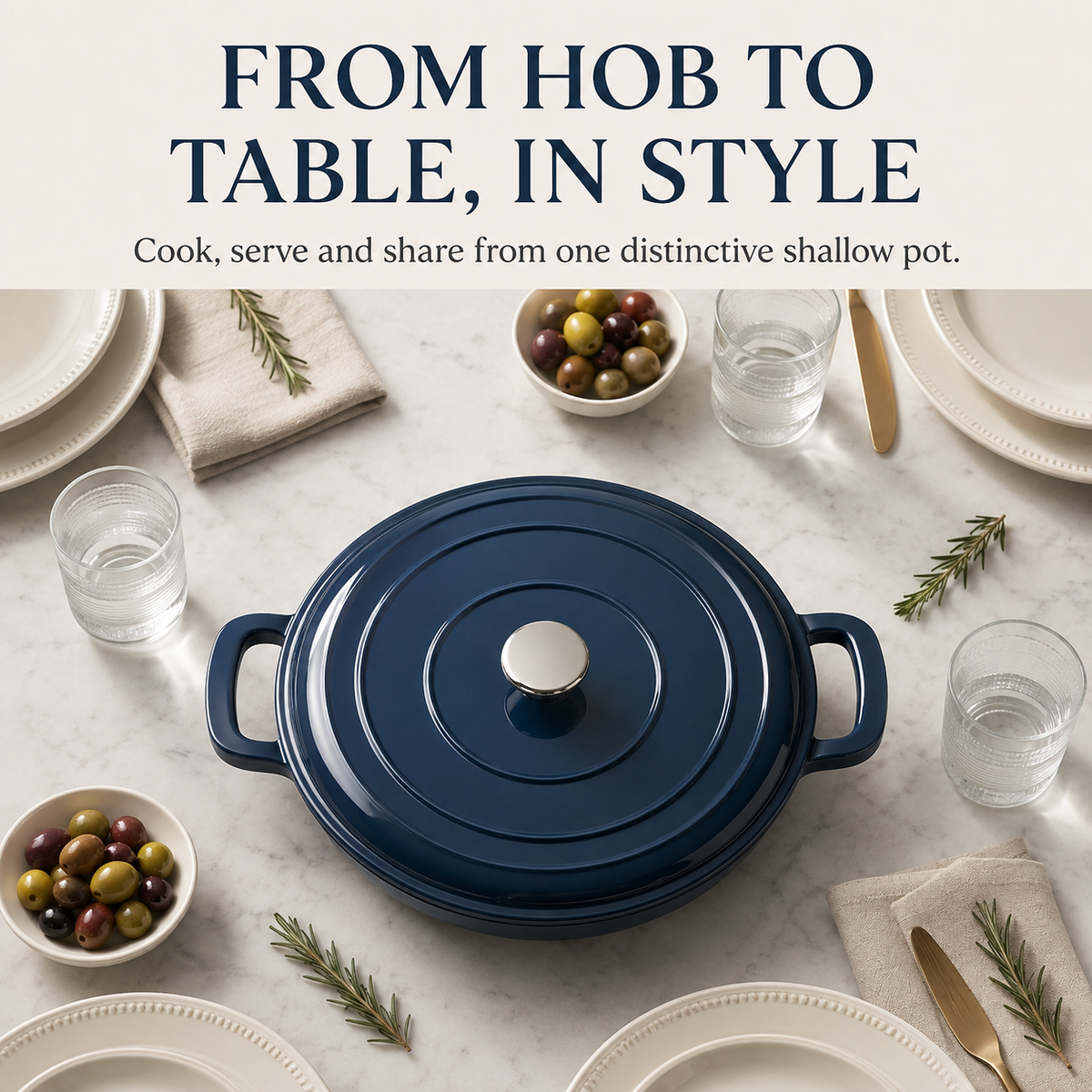 A navy blue enameled cast iron pot with a matching lid and side handles, styled on a white marble tabletop with plates, glasses of water, olives, and rosemary. Text at the top reads 'FROM HOB TO TABLE, IN STYLE'.